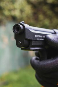 A detailed close-up of a gloved hand holding and aiming a semi-automatic pistol outdoors.
